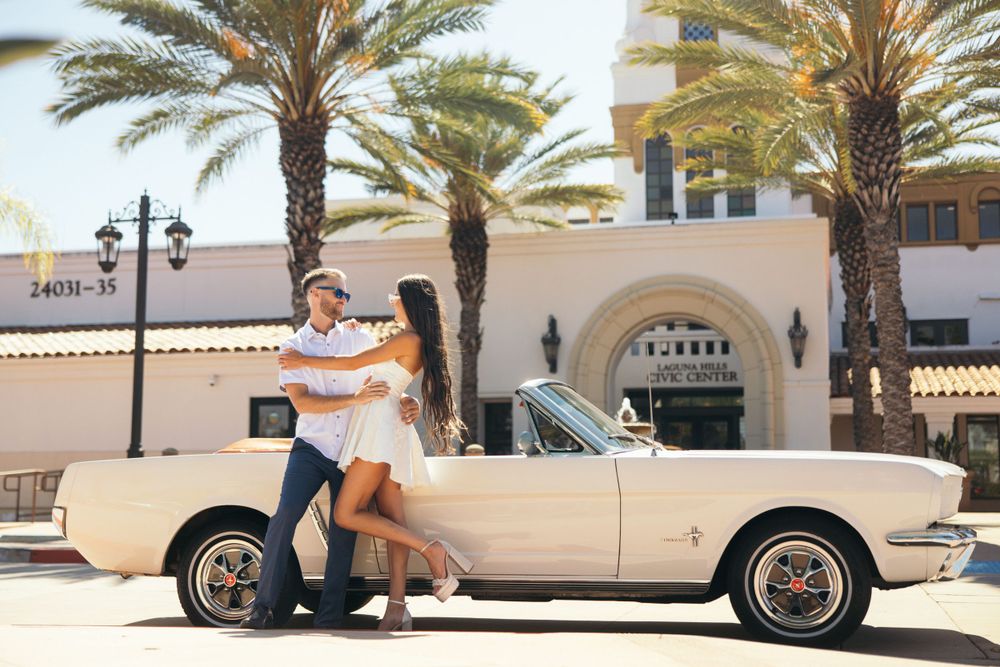 A romantic couple poses with a vintage cream-colored car in front of a Mediterranean-style building with palm trees.
