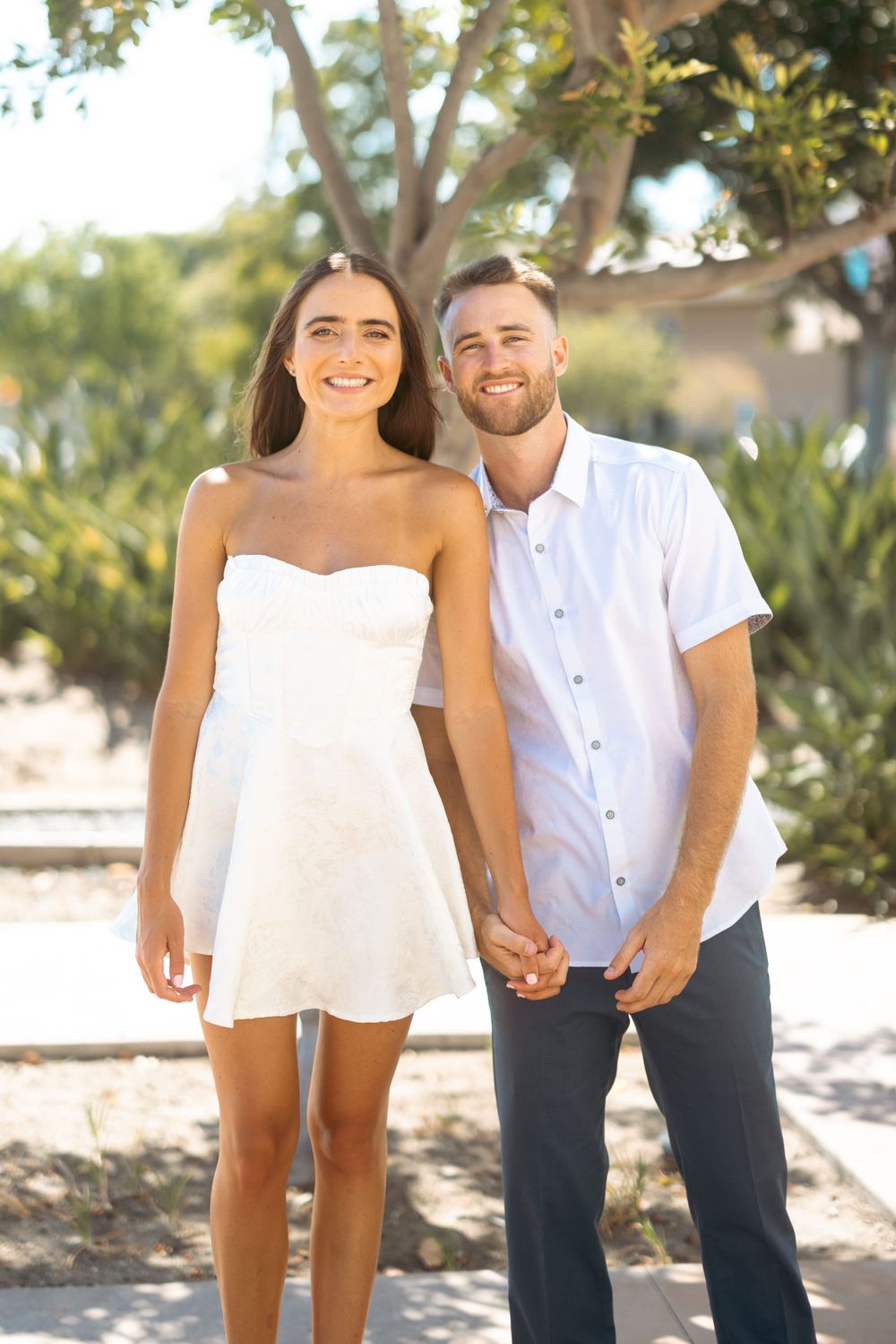 Couple poses together on sunny day against lush garden backdrop and white architecture.