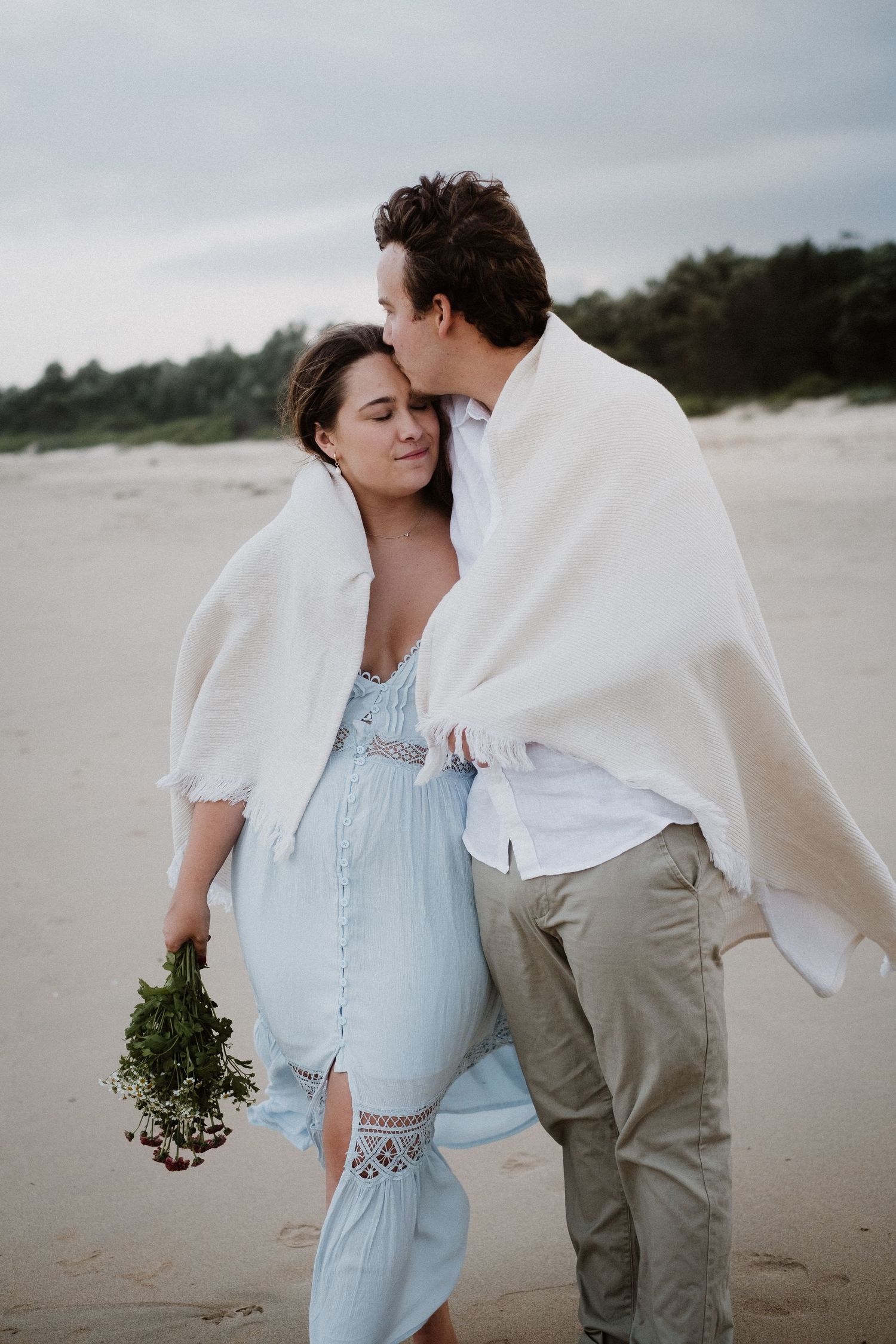 Romantic couple embraces on a cloudy beach while wearing light blue and beige outfits.