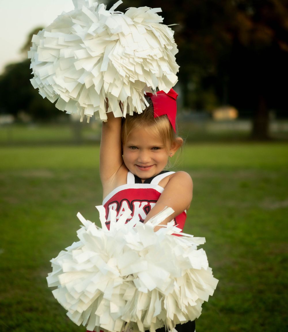Young cheerleader in red uniform holding white pom poms with an excited expression.