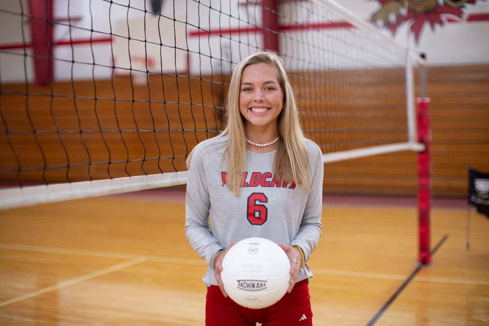 A volleyball player in a grey and red uniform poses with a ball in front of a net in an indoor gym.