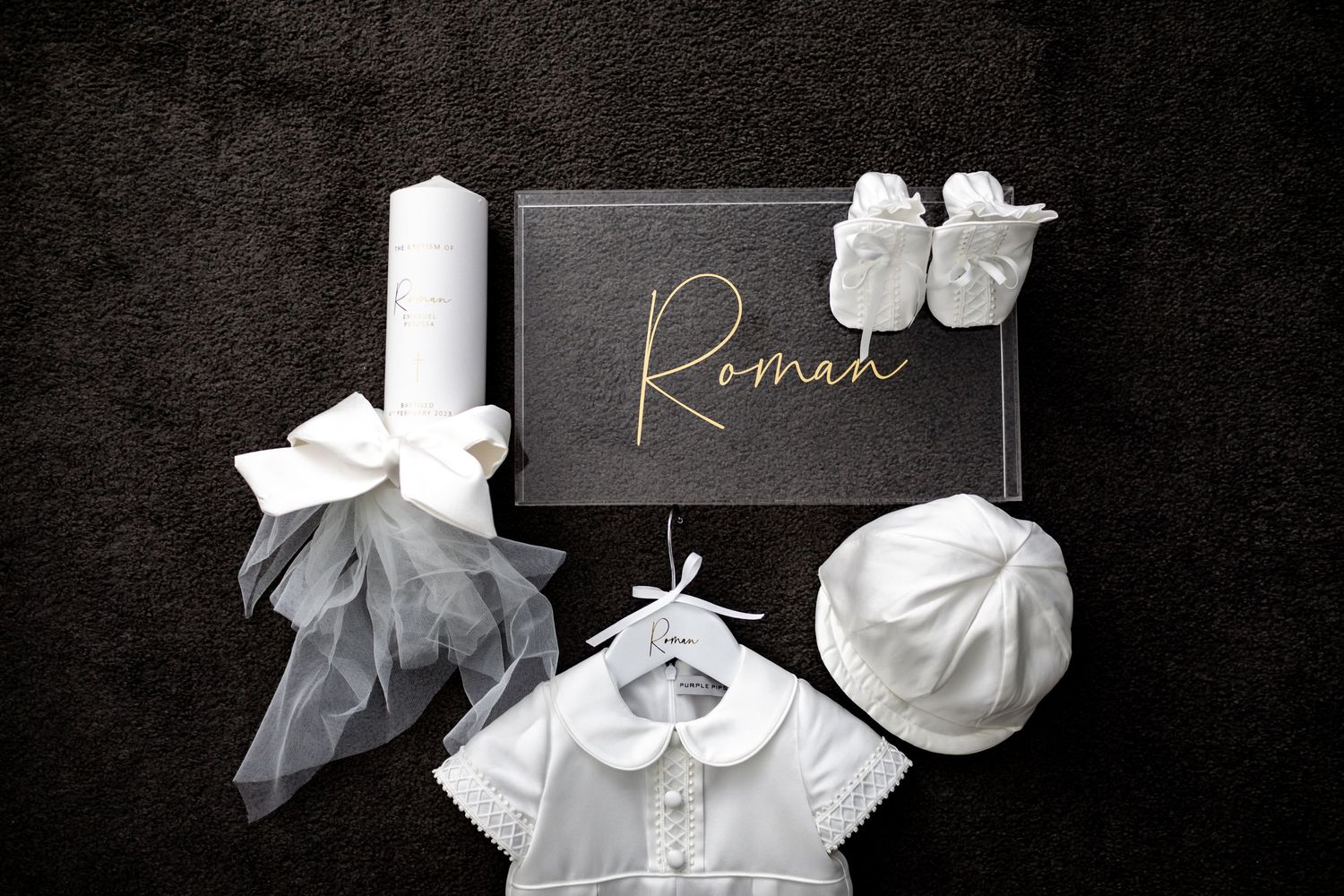 White baptism or christening accessories including candle, baby shoes, and outfit on dark background.