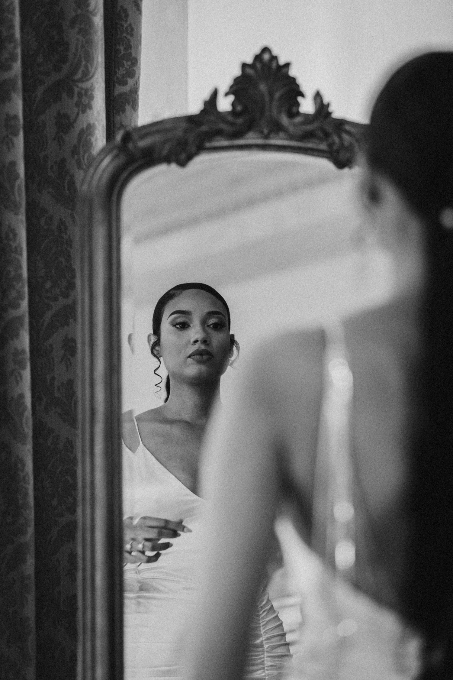 An elegant woman in a white dress looks at her reflection in an ornate vintage mirror.