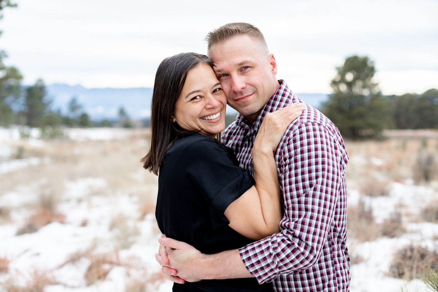 Husband and wife snuggle up and smile while standing in the snow in Black Forest, Colorado.