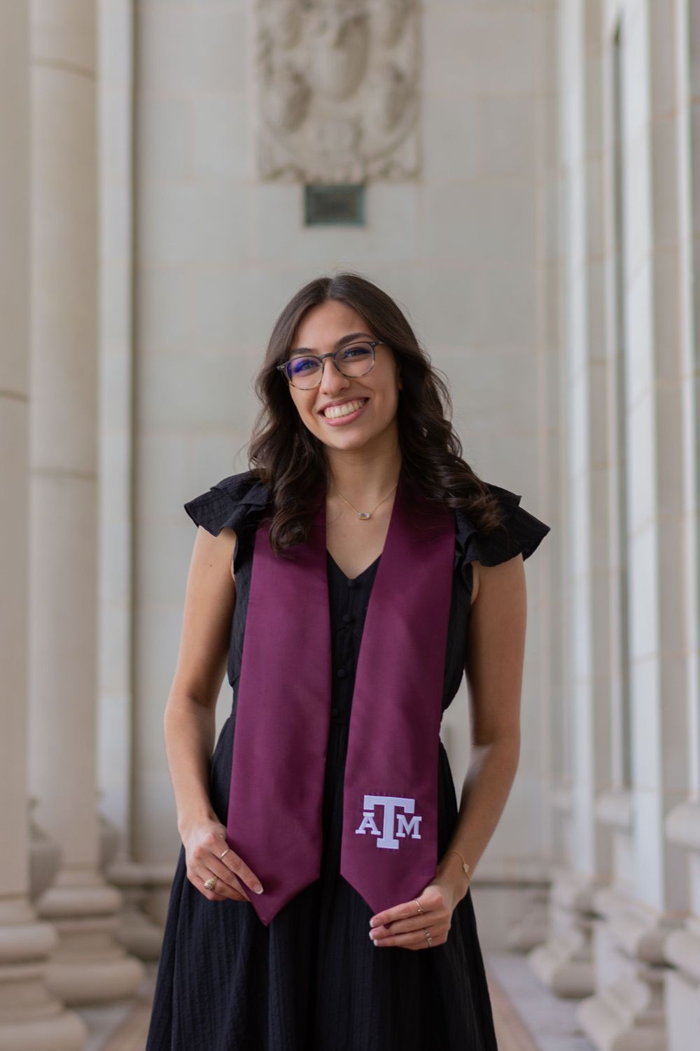 An Aggie graduating senior holds the bottoms of her maroon and white grad stole as she smiles. She's in a black dress at the Admin Building,