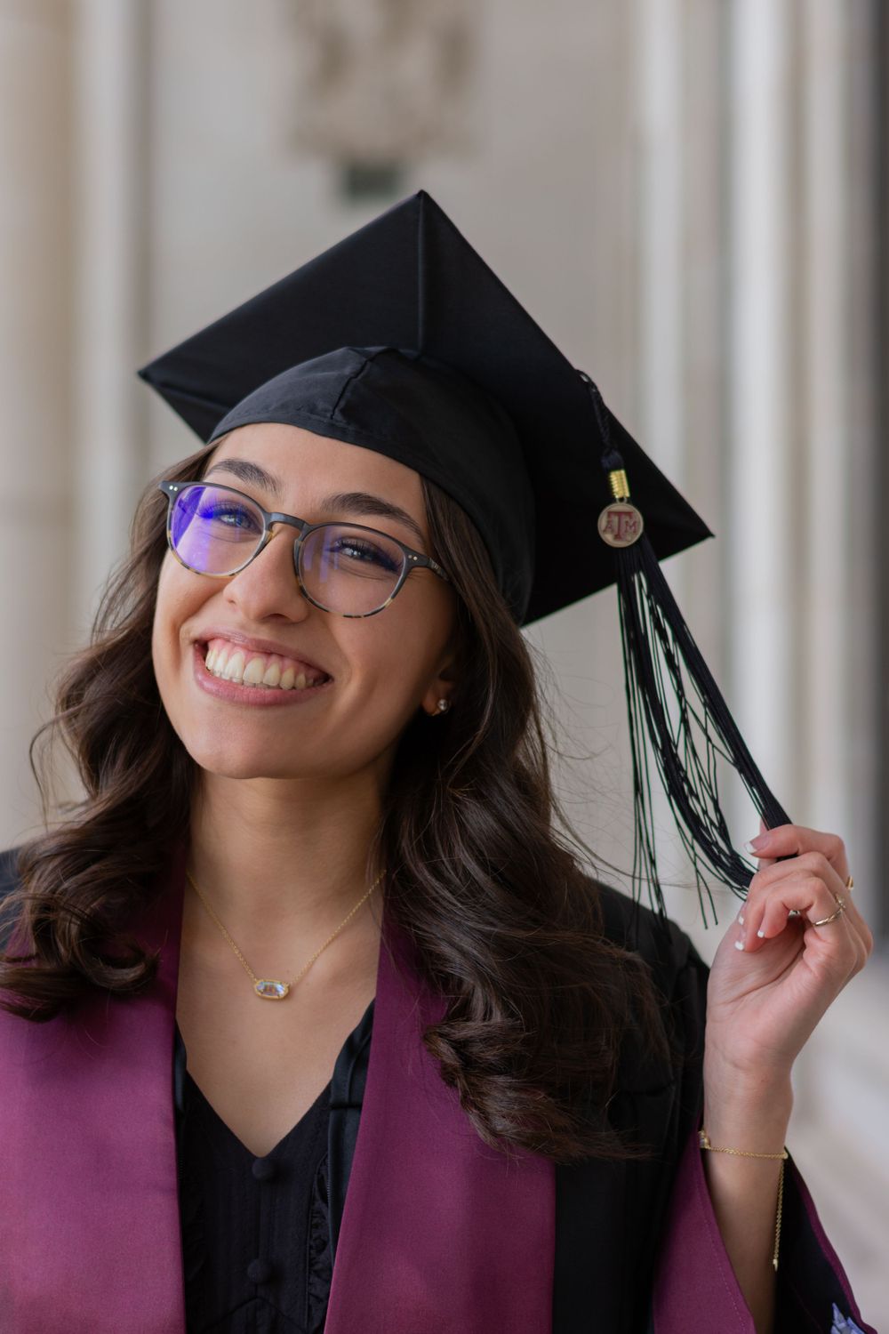 An Aggie grad wears her full graduation regalia over a black dress. She smiles while holding the strings of her grad cap's tassel.