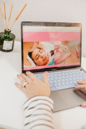 Hands typing on laptop keyboard with reed diffuser and beauty website displayed on screen.