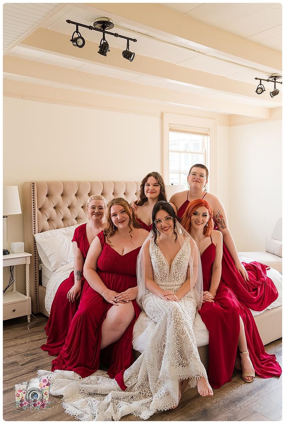 Bridal party in burgundy dresses gathered on bed with elegant decor and lighting.