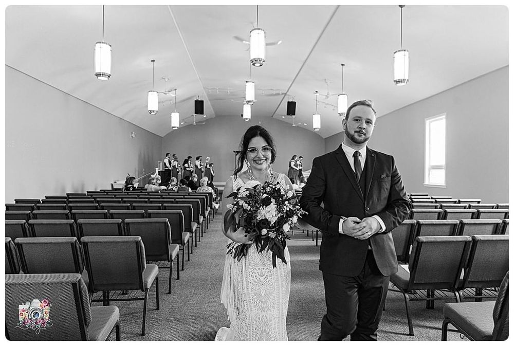 Black and white photo of couple walking down church aisle with pendant lights overhead.