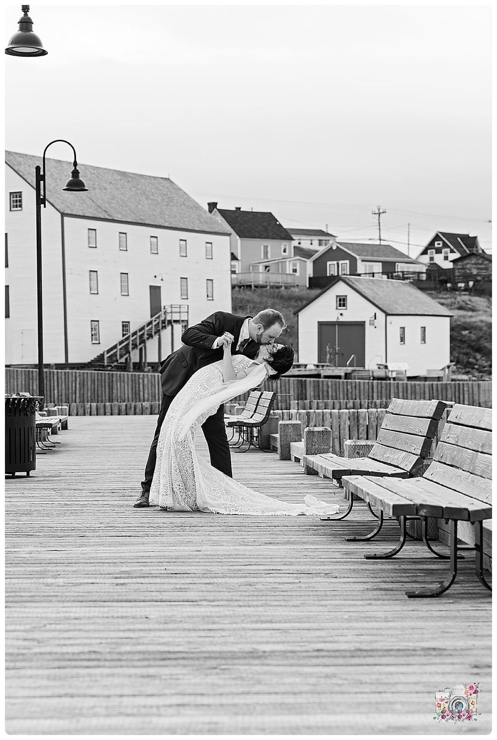 Black and white photo of a romantic dip pose on a wooden boardwalk with coastal buildings.