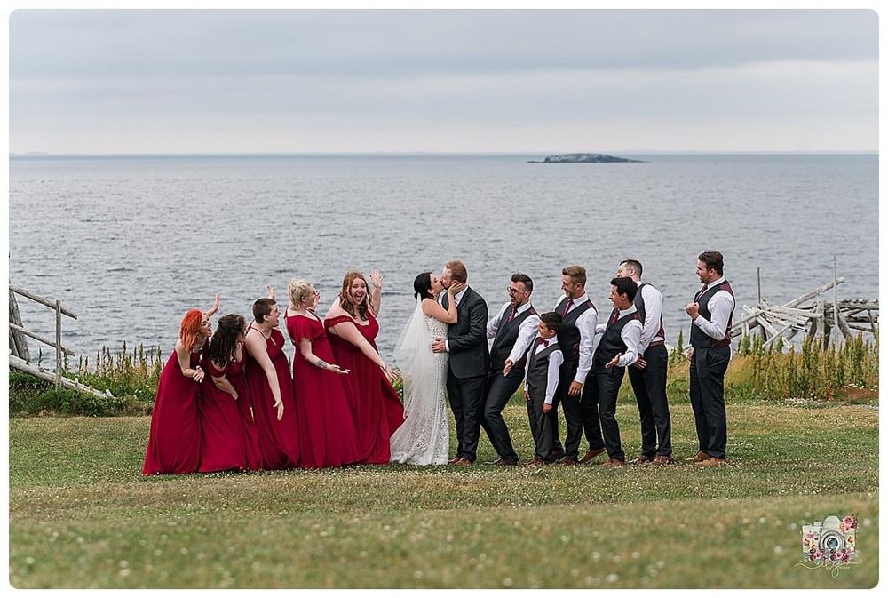 Wedding party in red and black attire poses playfully on grassy cliff overlooking the ocean.