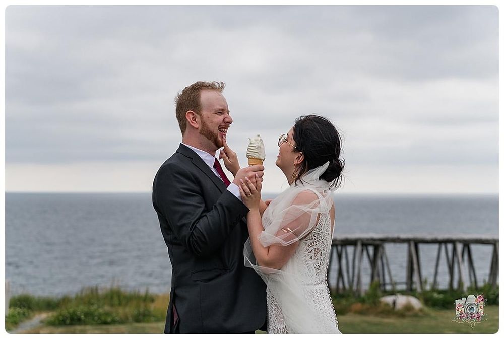 Couple shares ice cream cones by the ocean with wooden pier structure in background.