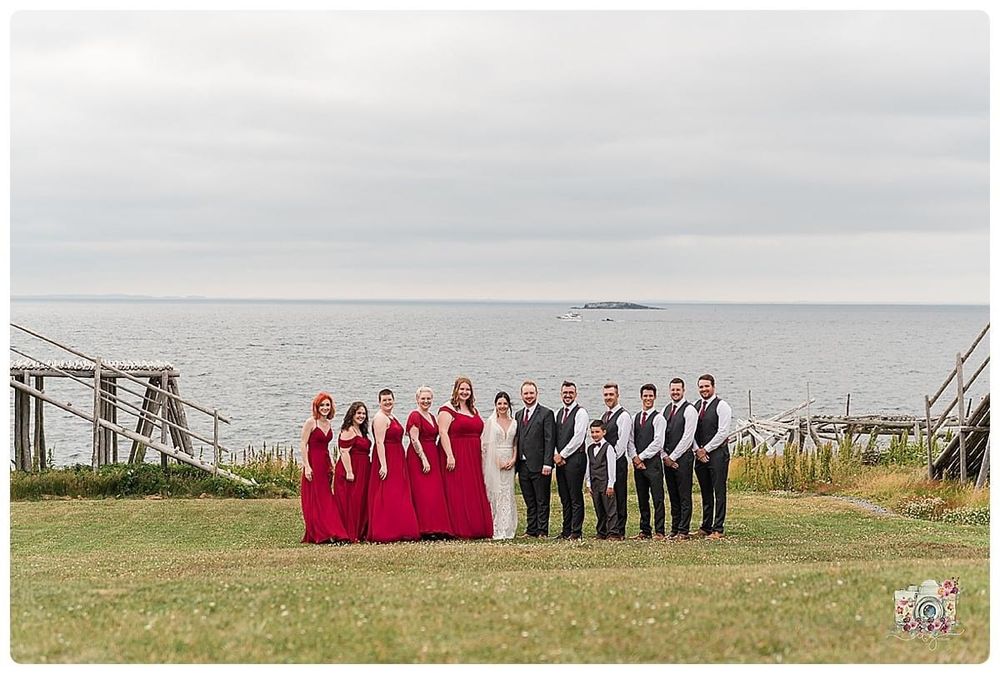 Wedding party in red and black attire poses on oceanside cliff with wooden structures.