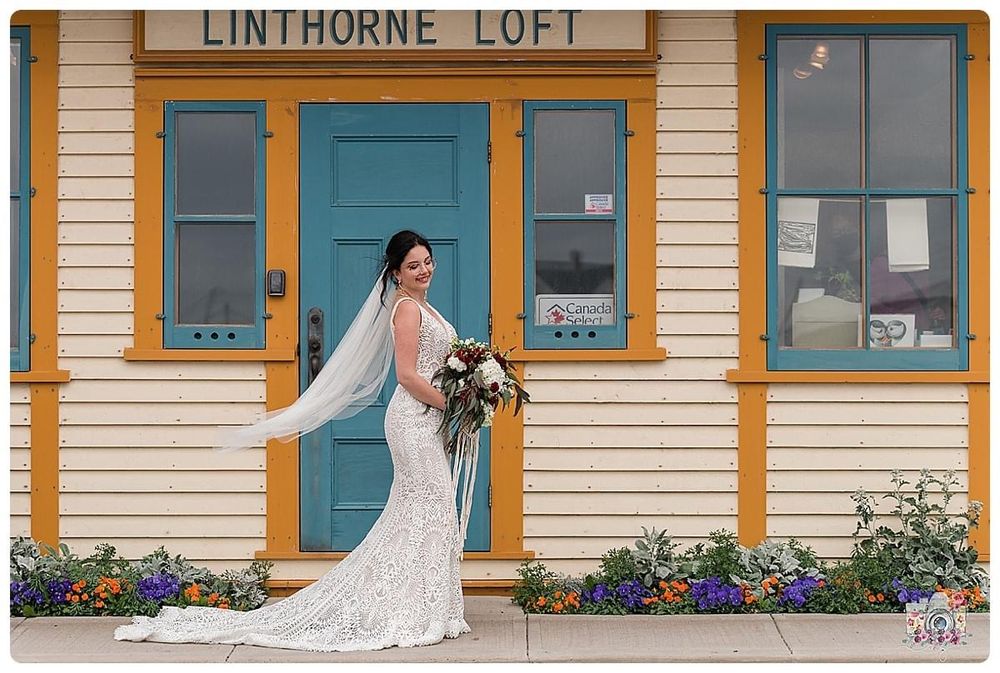 Bride in lace dress poses solo in front of yellow Linthorne Loft building with flowers.