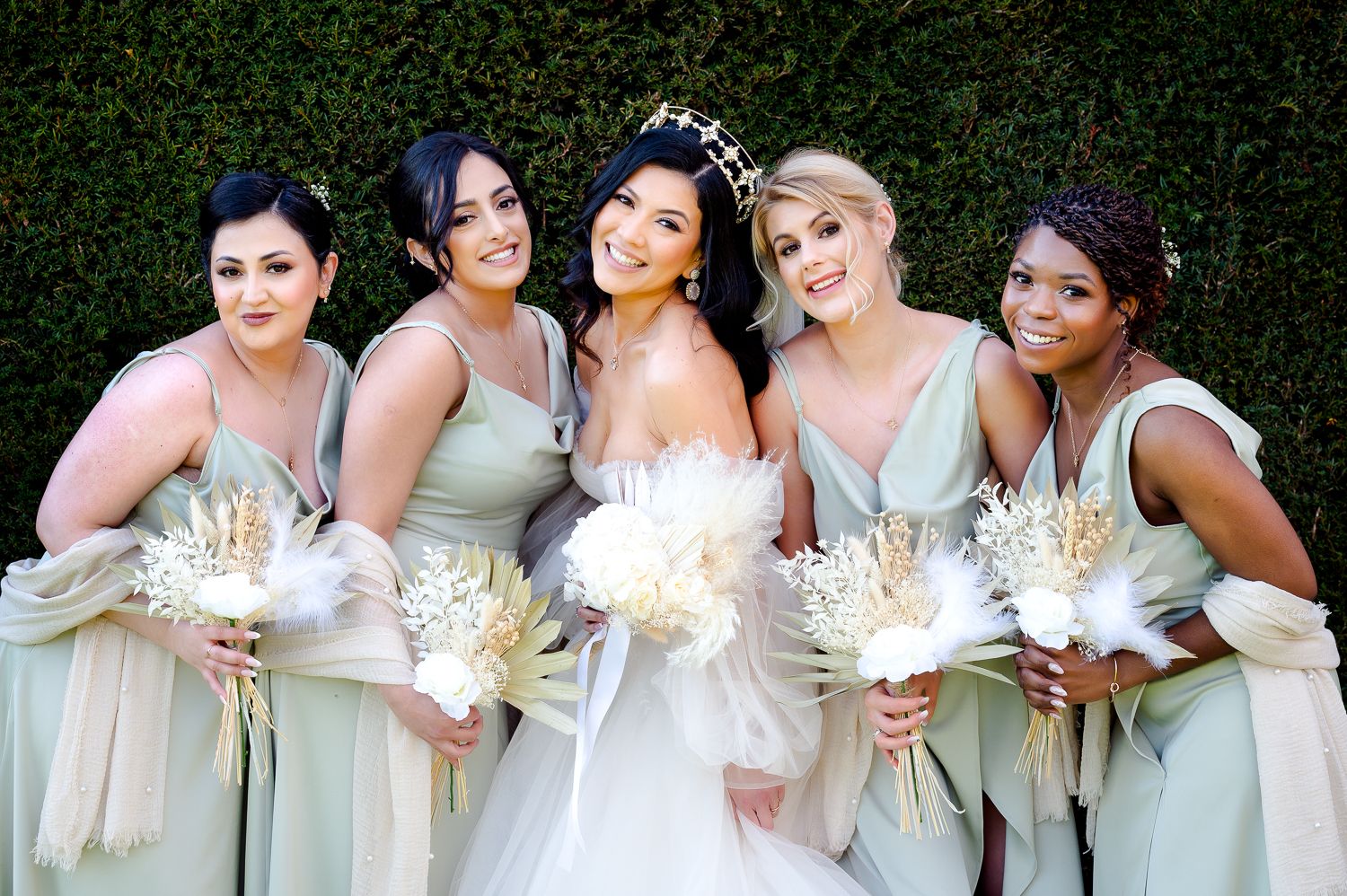 A bridal party in pale colored dresses holds white bouquets while posing against a green hedge backdrop.
