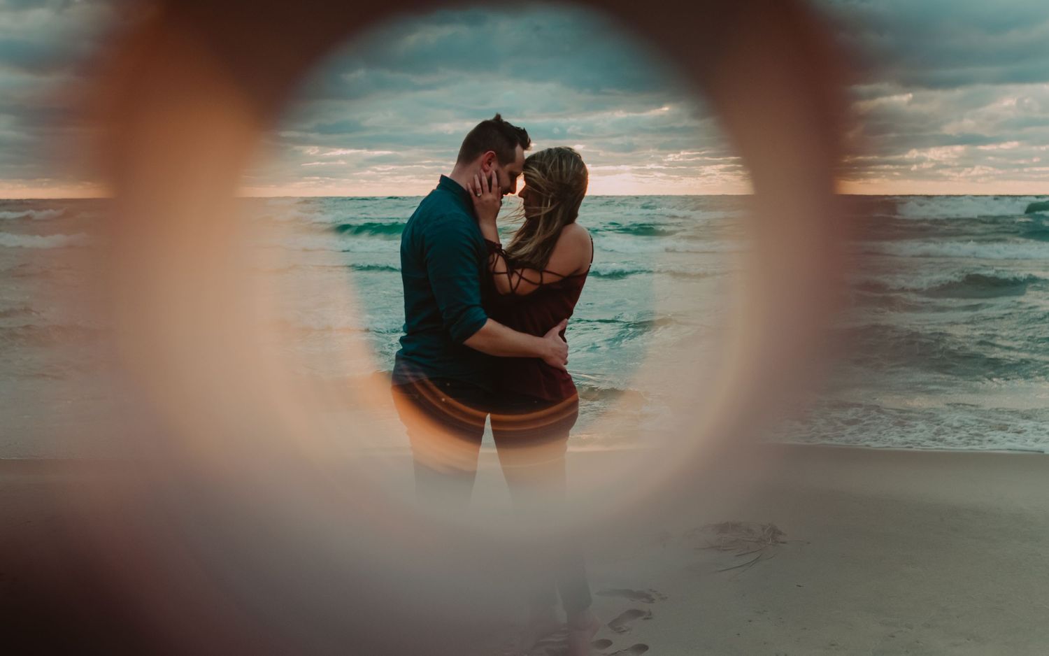 A couple embraces on a beach at sunset, framed through a circular foreground element.