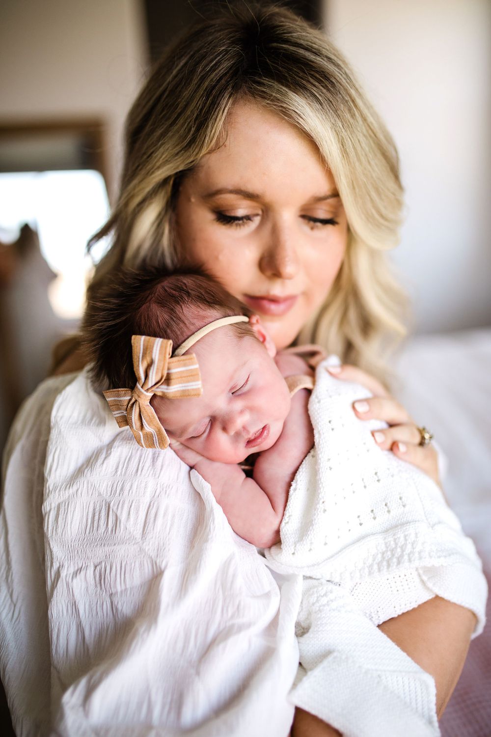 Blonde mother tenderly holds sleeping newborn baby girl wearing striped bow headband wrapped in white swaddle.