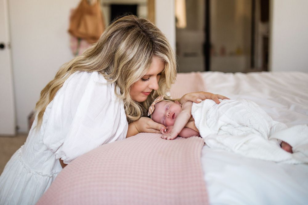 New mother in white dress leans over bed to kiss her sleeping newborn baby wrapped in white blanket.