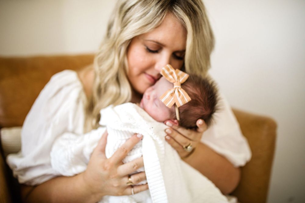 Blonde mother closes eyes while gently nuzzling her swaddled newborn baby with striped bow headband.