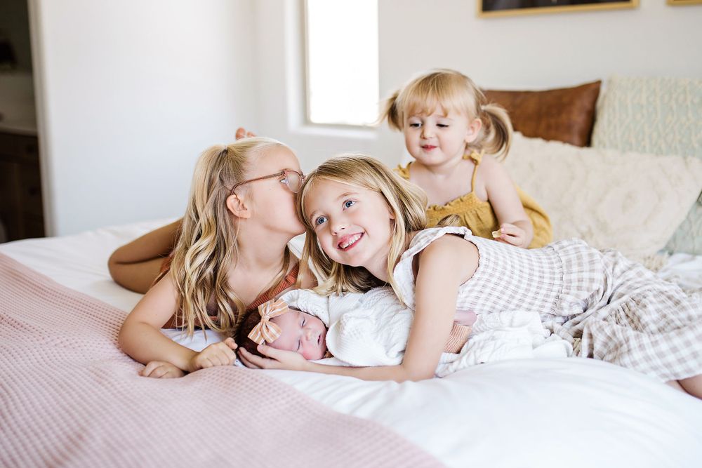 Three sisters cuddle together on pink bedspread with newborn baby, one giving a sweet kiss.