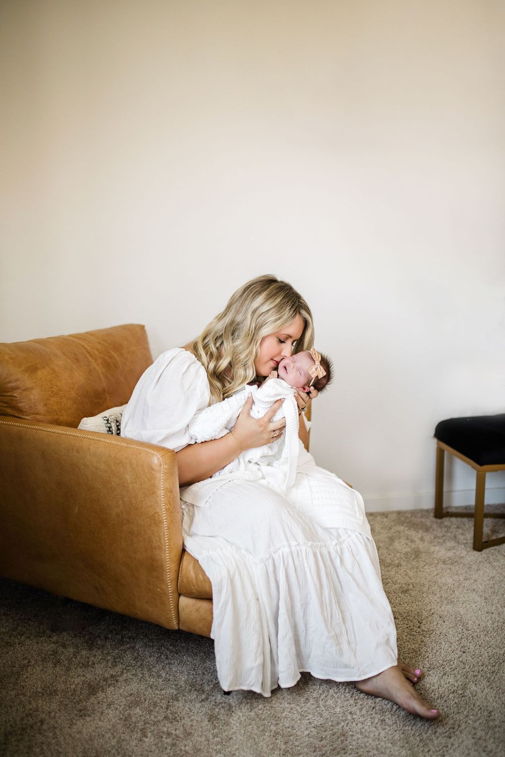 New mother in white dress sits on tan leather sofa kissing her newborn baby girl tenderly.