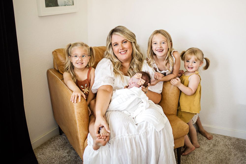 Happy mother in white dress poses on leather sofa with her newborn and three young daughters.