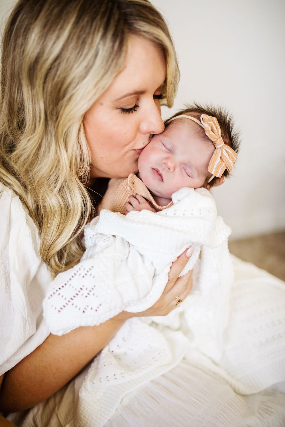 Loving mother kisses the cheek of her sleeping newborn baby girl wearing a peach striped bow.