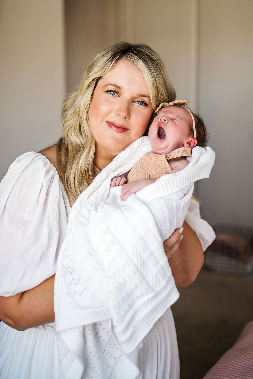 Smiling blonde mother holds her yawning newborn baby wrapped in white blanket.