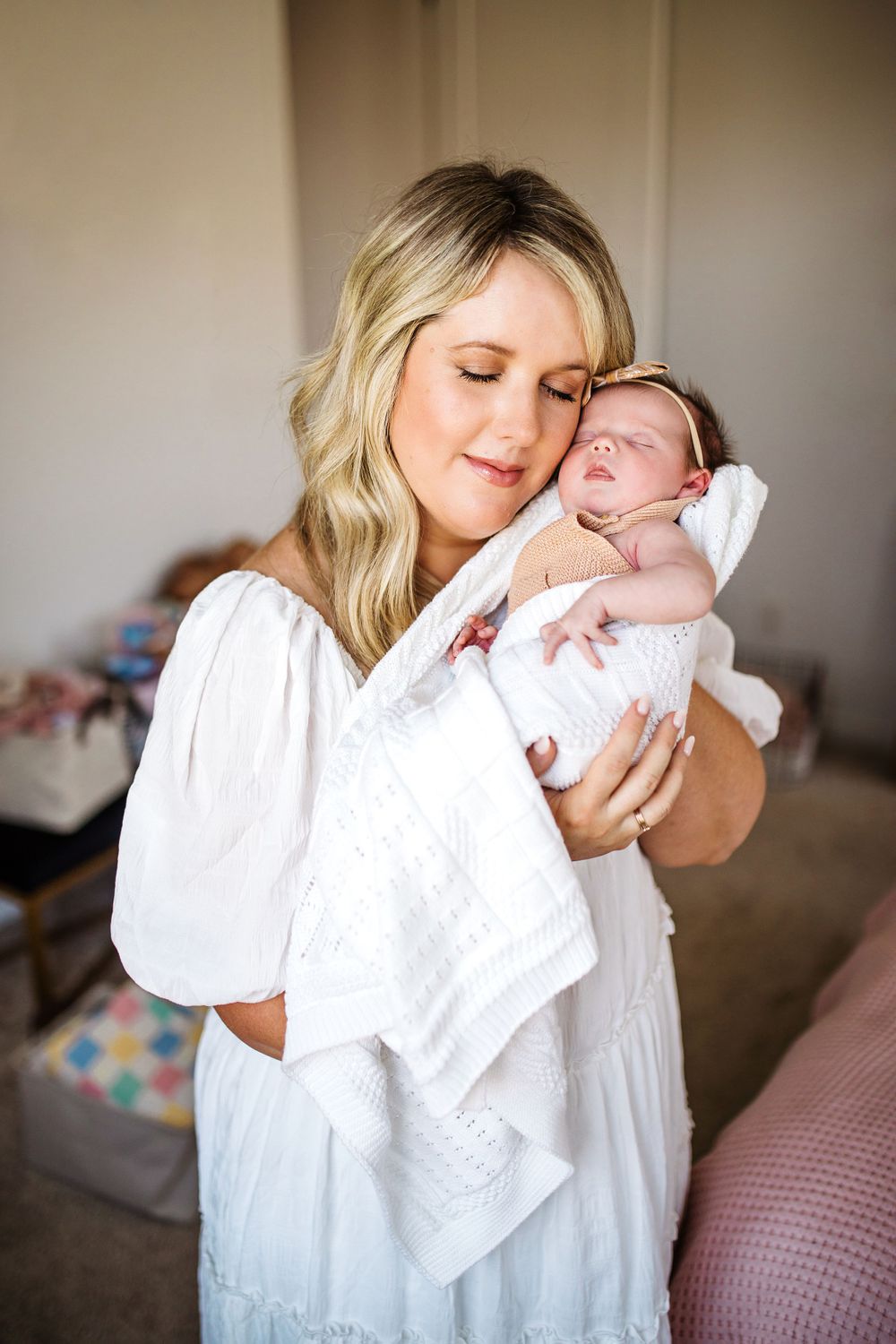 Tender moment as blonde mother cuddles her sleeping newborn wrapped in white.