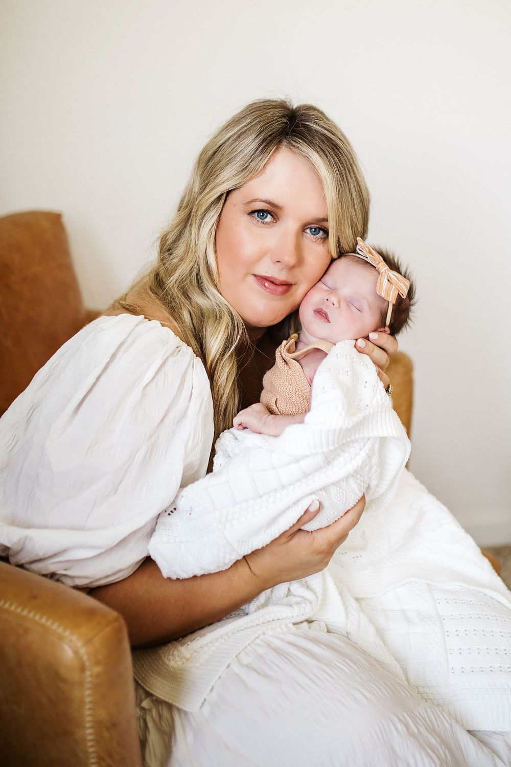 Smiling blonde mother with blue eyes cuddles sleeping newborn baby girl in white swaddle on leather chair.
