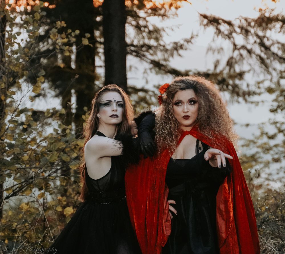 Two models in gothic-inspired outfits pose dramatically in a moody autumn forest setting.