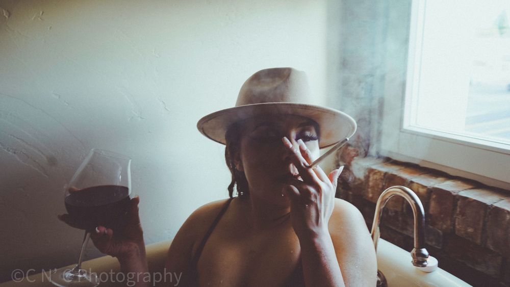 Atmospheric photo of someone in a fedora hat smoking by a window in moody lighting.