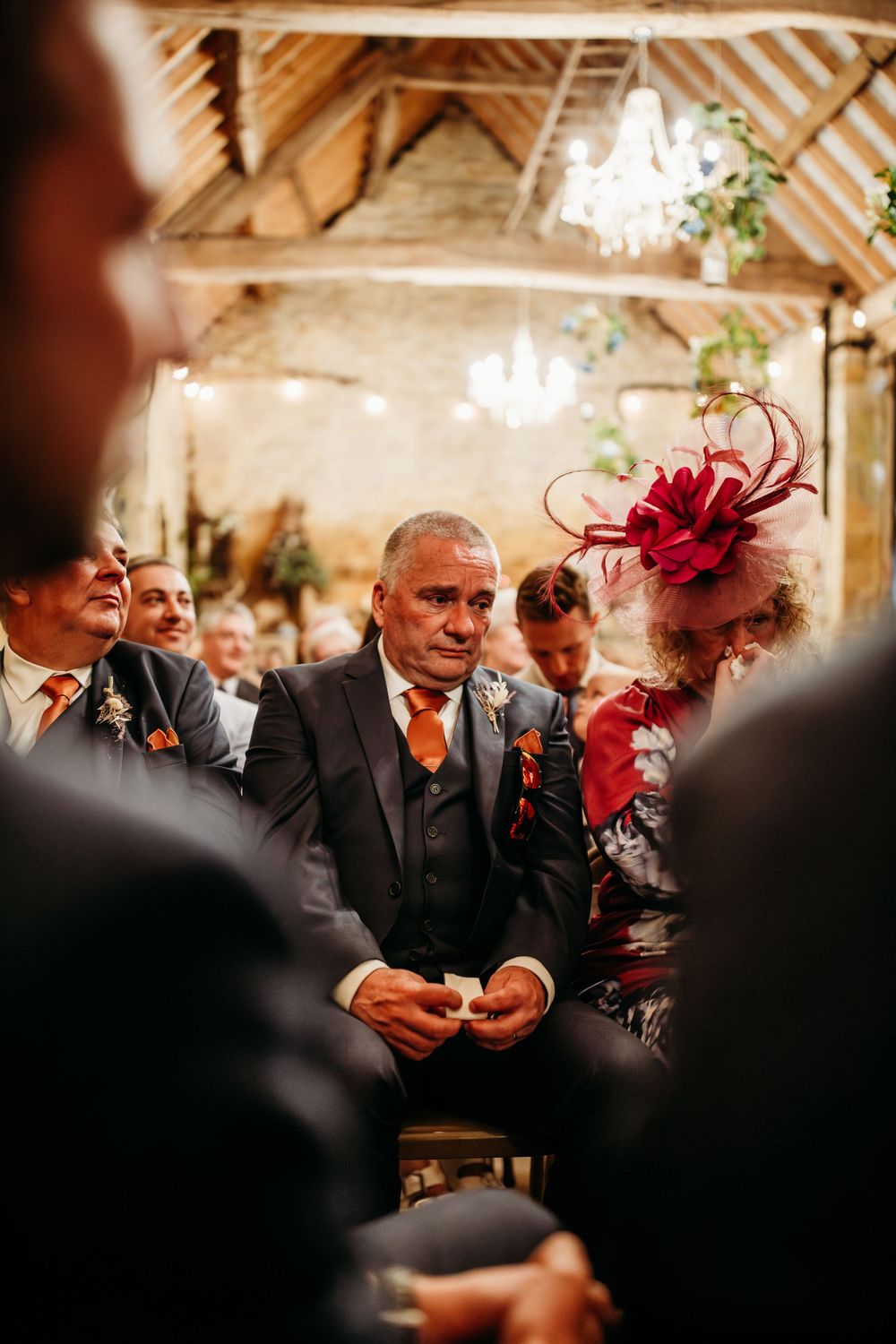 Wedding guests in formal attire gather beneath wooden beams and chandeliers during ceremony.