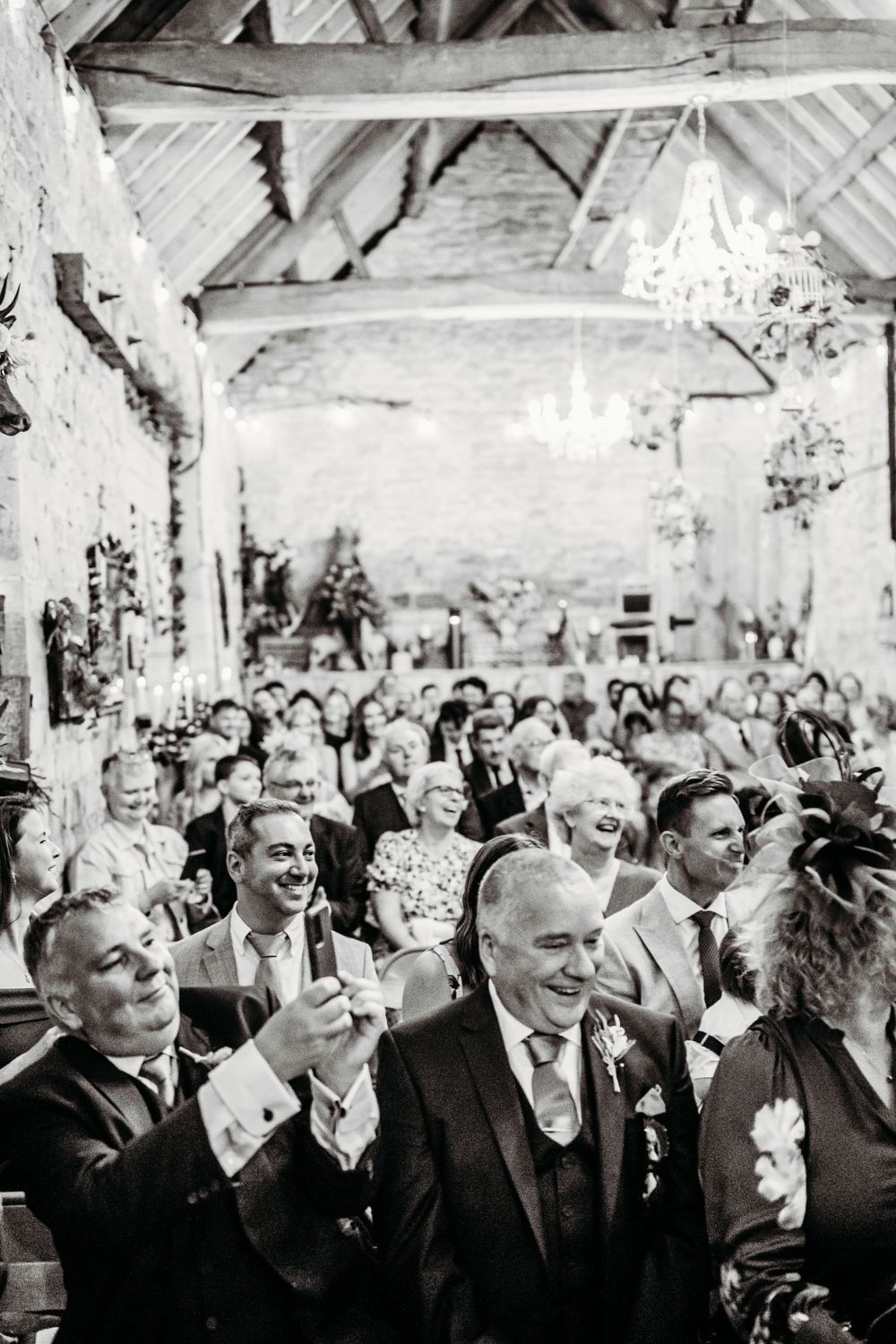 Black and white photo of wedding guests enjoying a celebration in a rustic venue with chandeliers.
