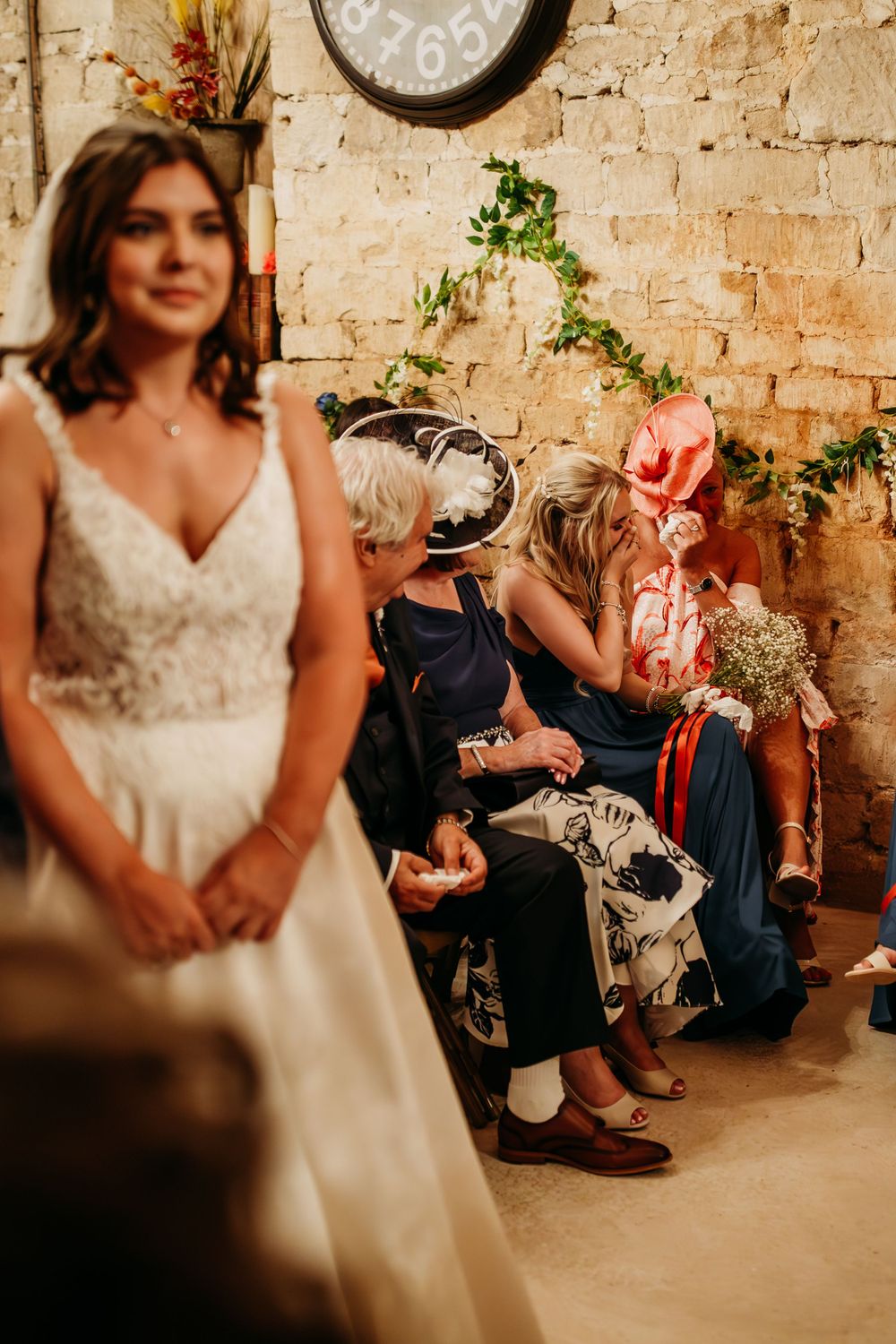 A bride in a white lace gown stands near exposed brick walls decorated with greenery.