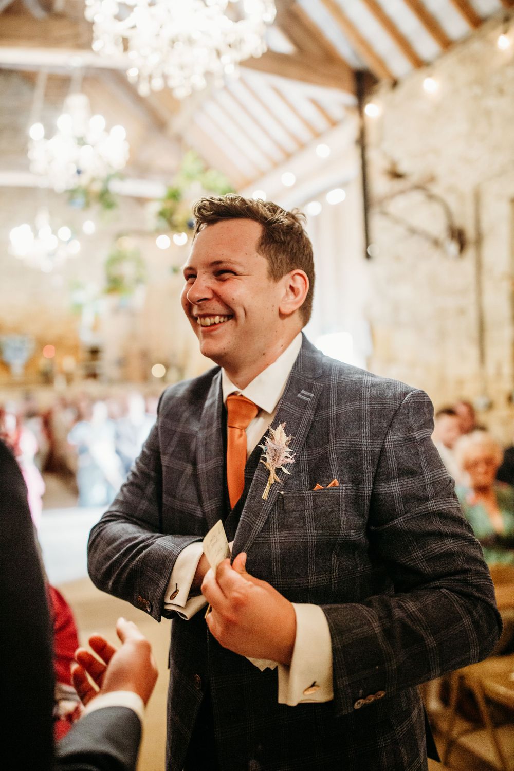 A groom in a checkered suit and orange tie smiles during his wedding celebration.