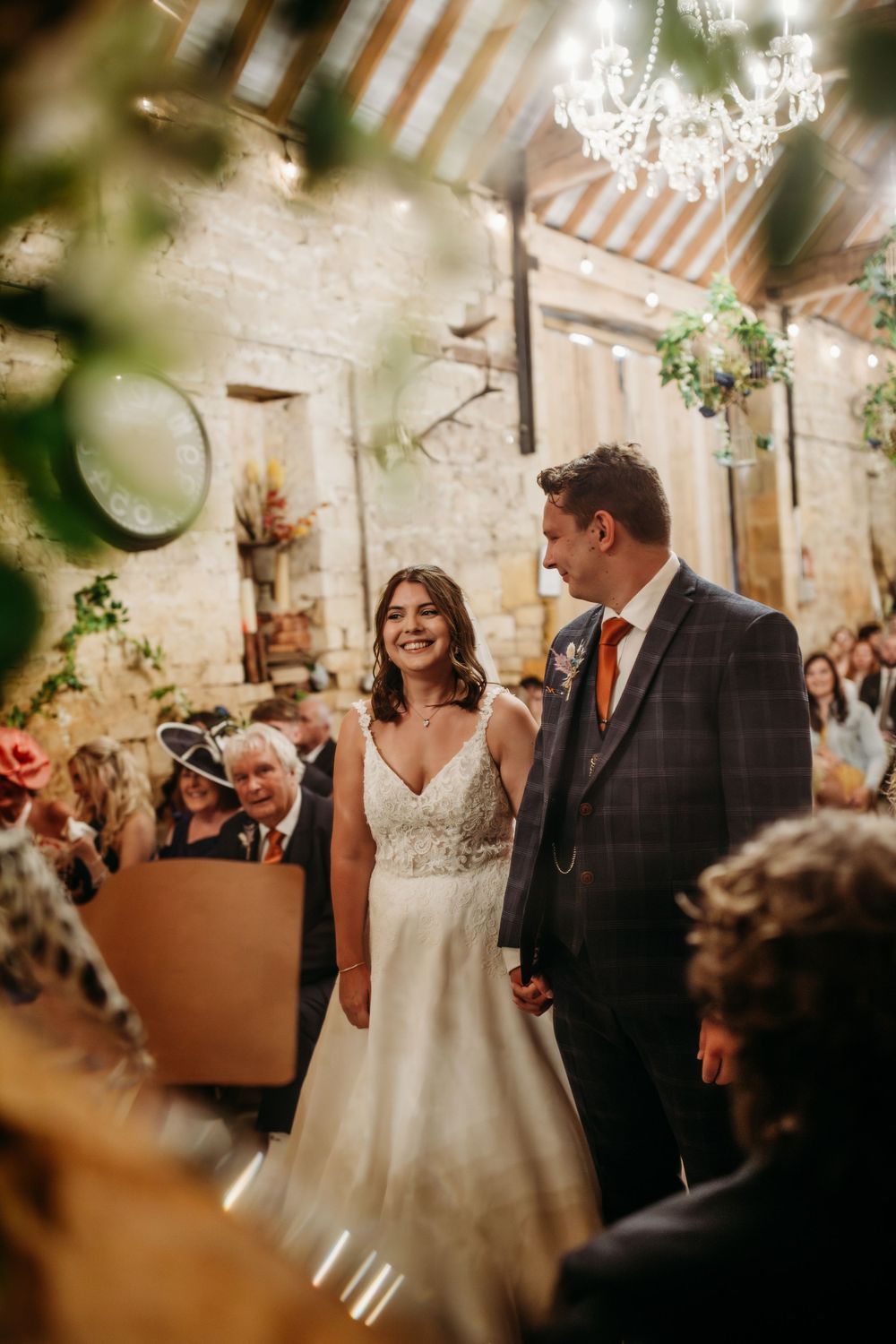 Couple stands together during wedding ceremony in stone-walled venue with chandelier and greenery.