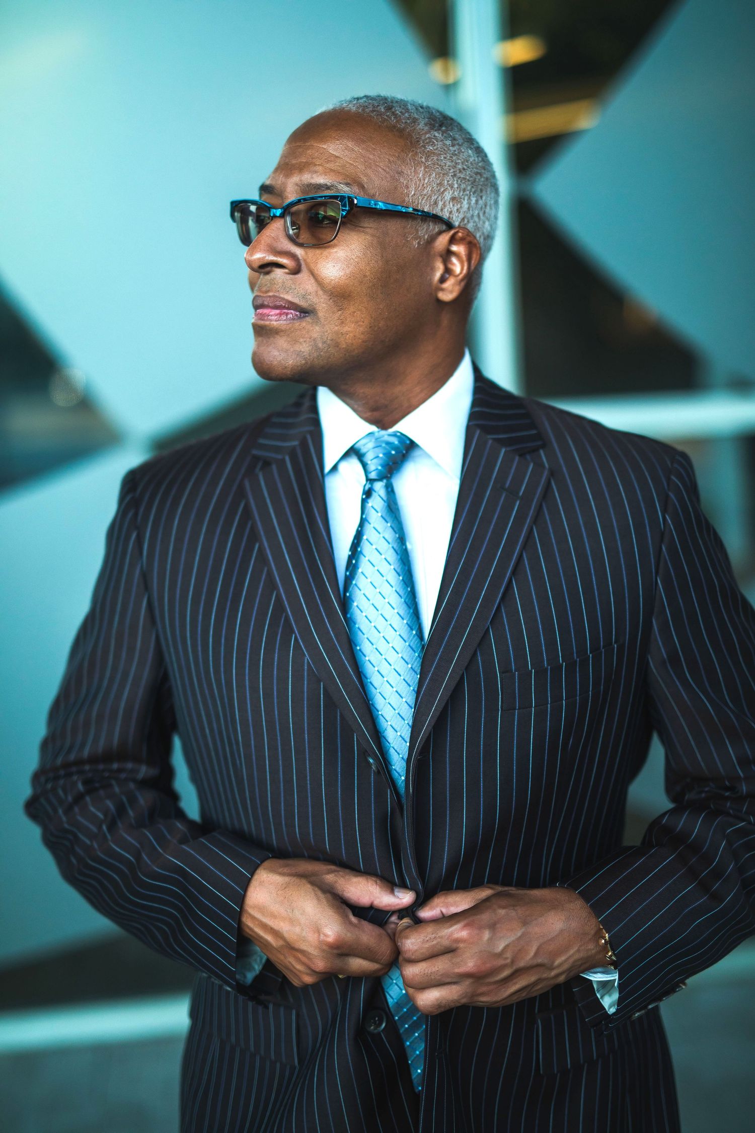 Professional in navy pinstripe suit with blue tie and glasses stands confidently against modern backdrop.