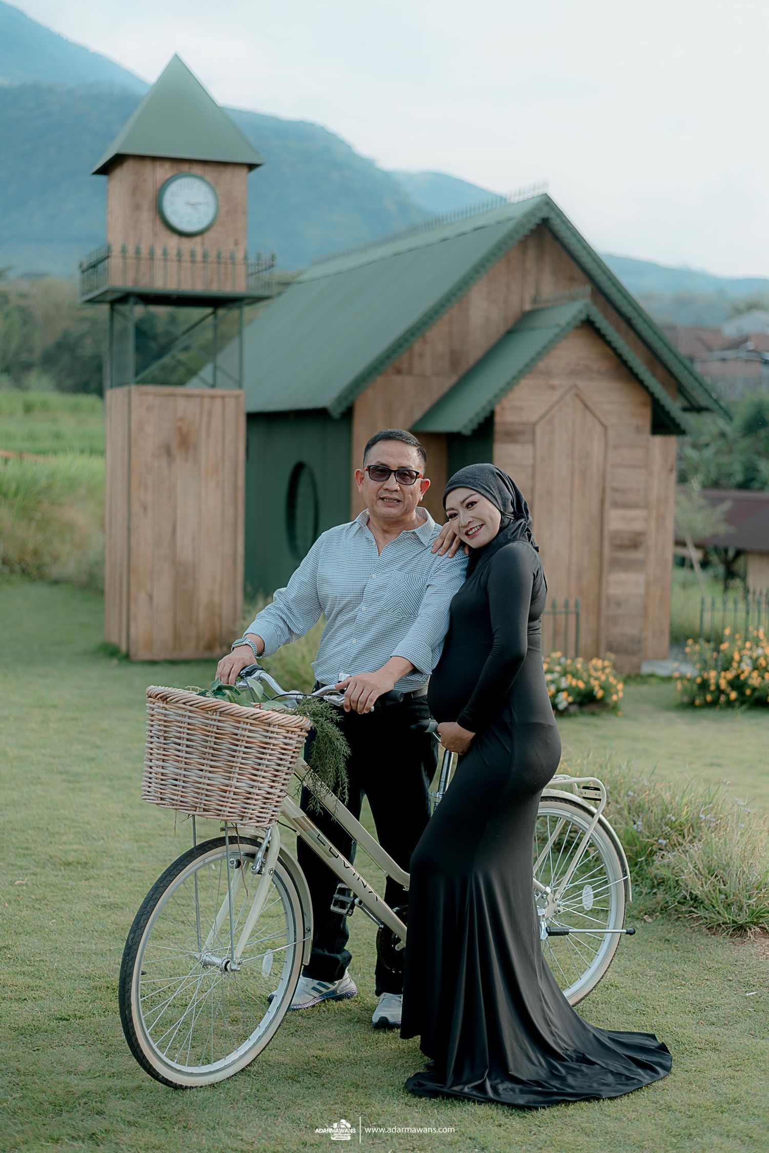 Couple sharing intimate moment with bicycle near wooden chapel and clock tower in mountain landscape.