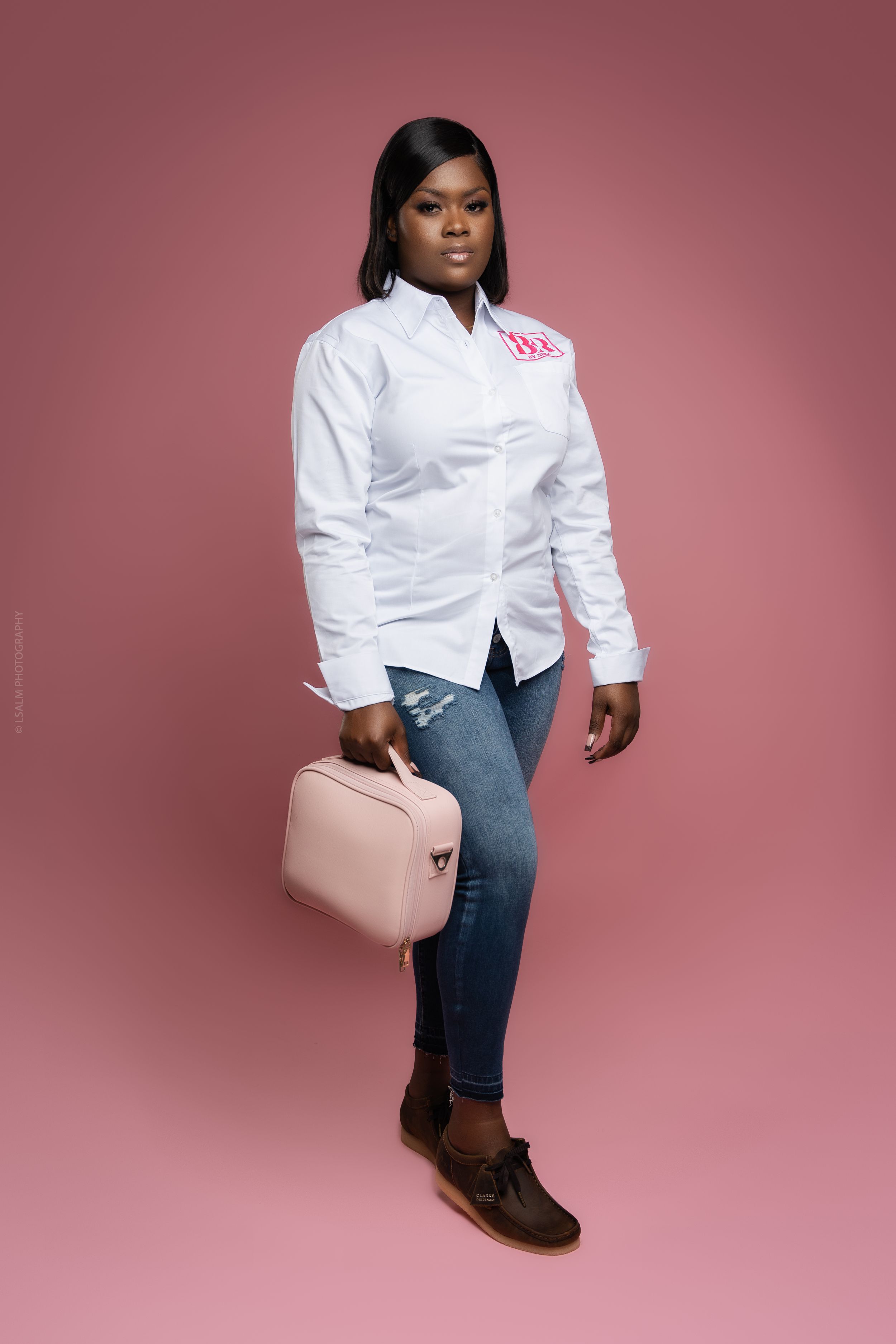 Professional wearing a crisp white button-down shirt, blue jeans, and brown shoes holds a pink handbag against pink backdrop.