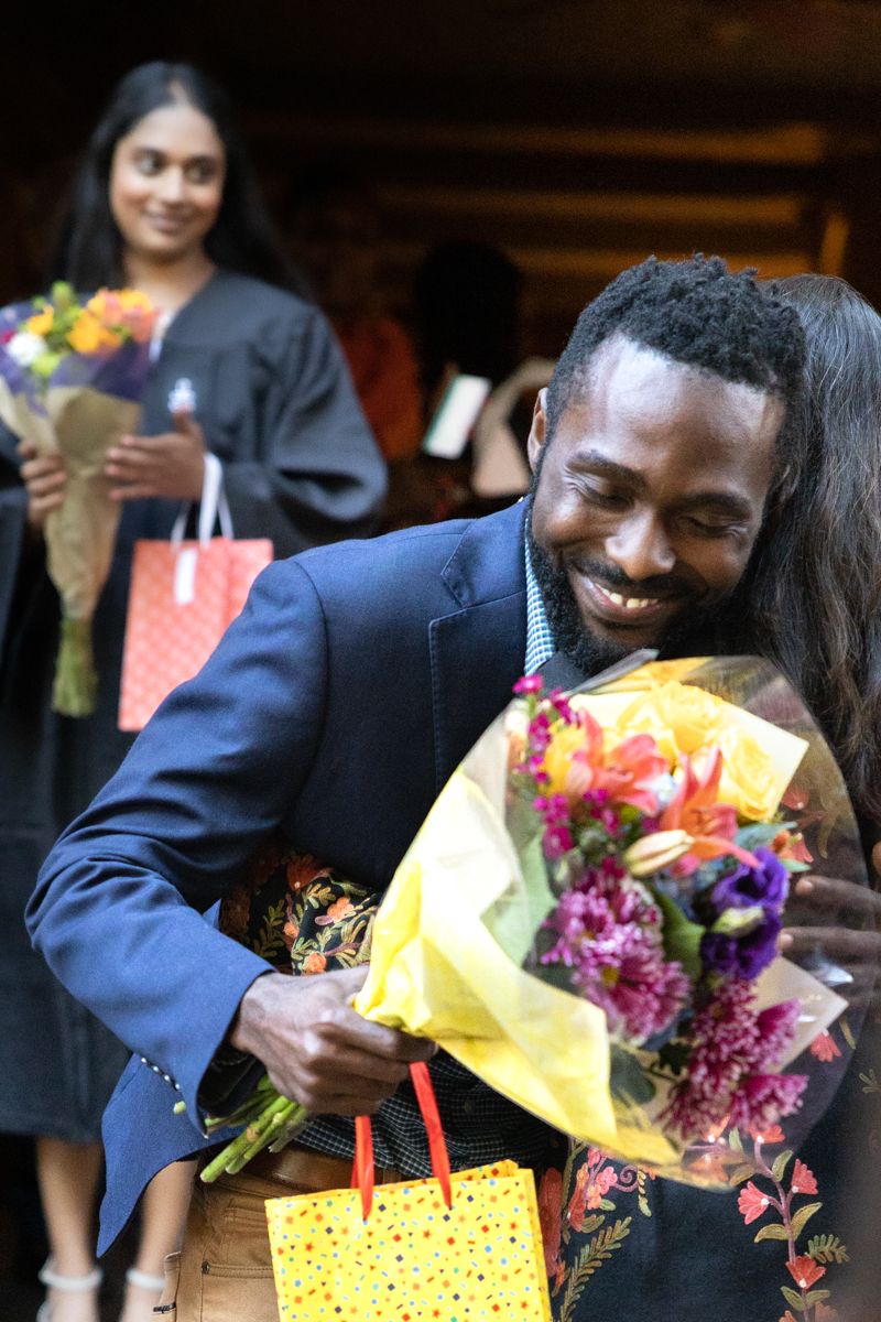 A man and woman embrace at a celebration. The man, who is Black, holds flowers and smiles while a third woman looks on