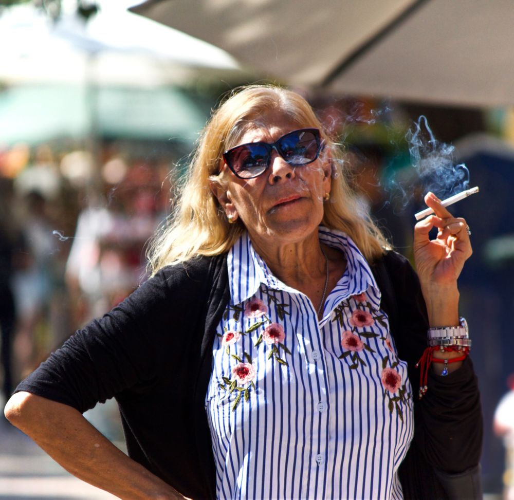 A blonde woman in sunglasses holds a cigarette in her left hand, off which smoke curls. Her hand is on her hip
