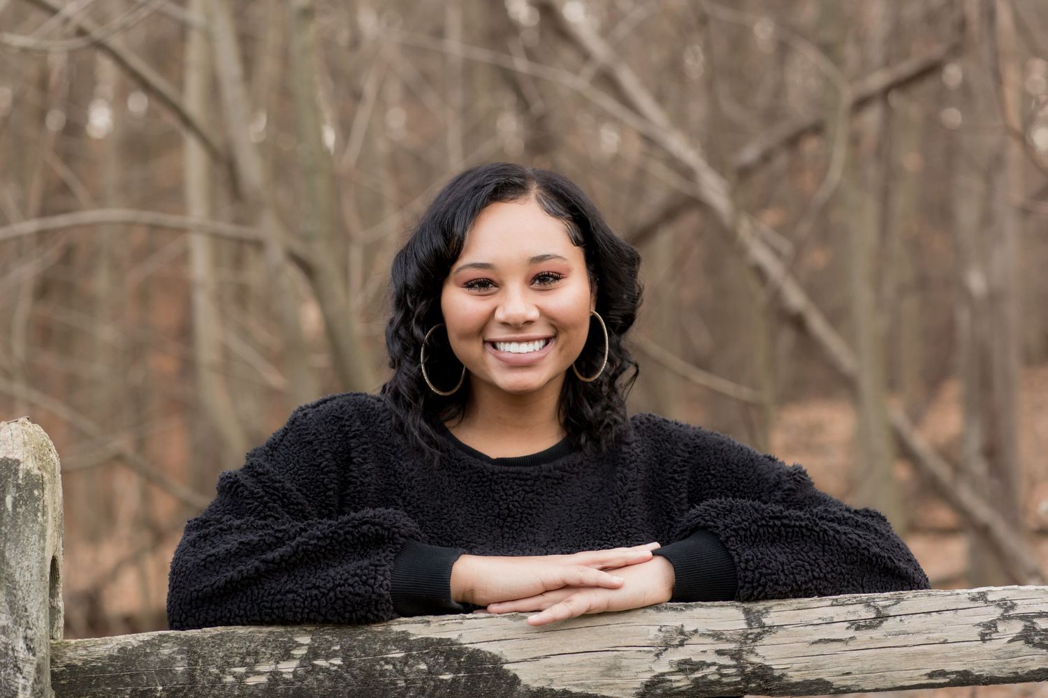 Smiling young woman in black sweater leaning on stone wall in woods.