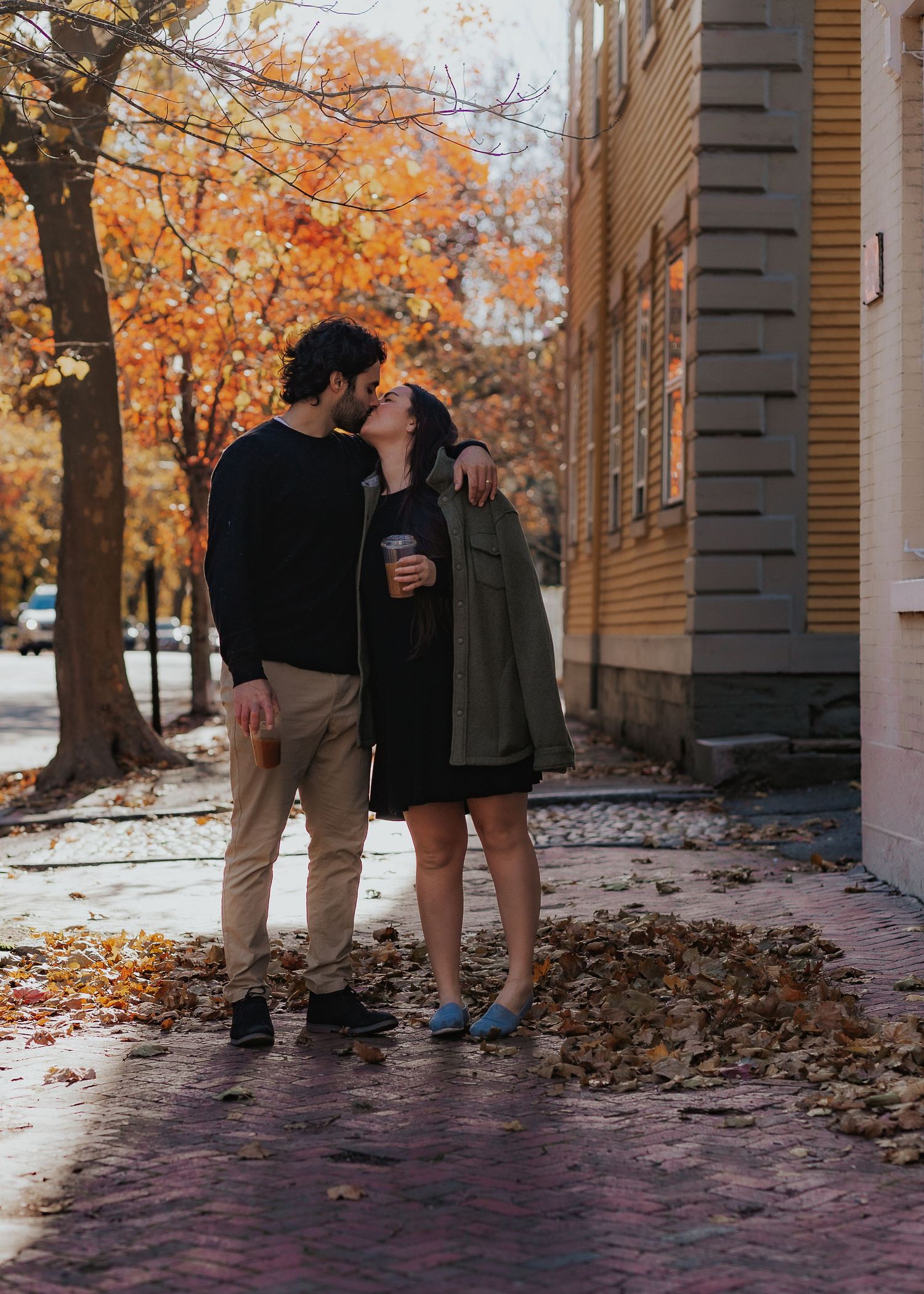 A couple stands together on a leaf-covered walkway between columns in Providence New England during autumn with trees showing fall colors.