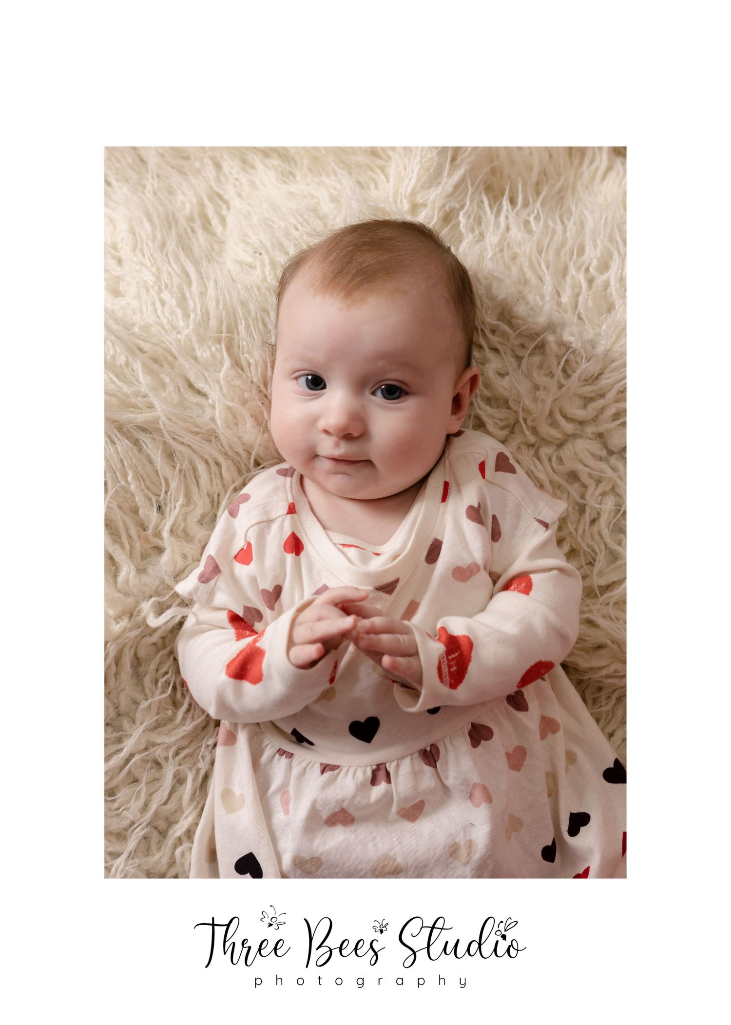 Baby in heart print outfit lying on fluffy cream rug during portrait session.