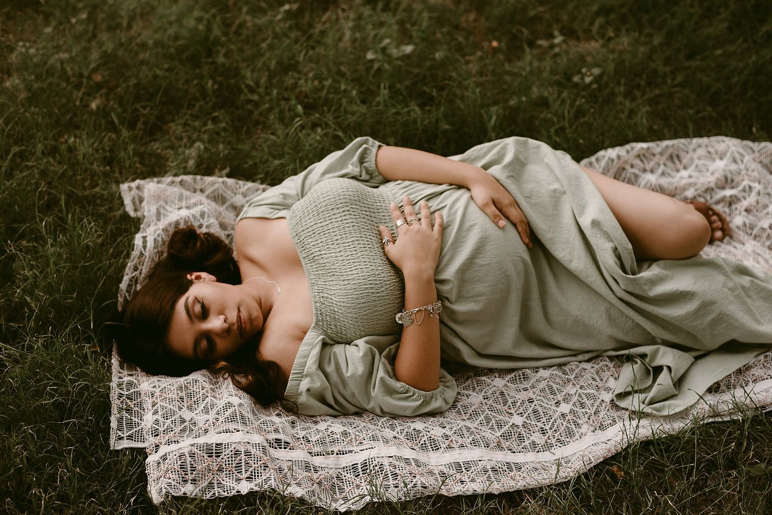 A maternity photo on a white lace blanket outdoors shows a woman in soft gray dress resting peacefully.