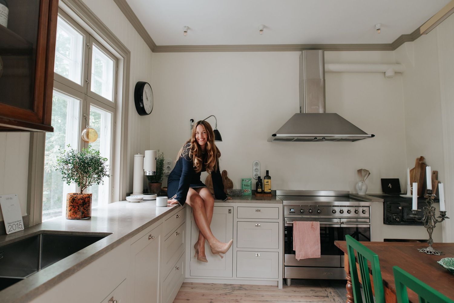 Woman relaxing on modern white kitchen counter next to large windows.