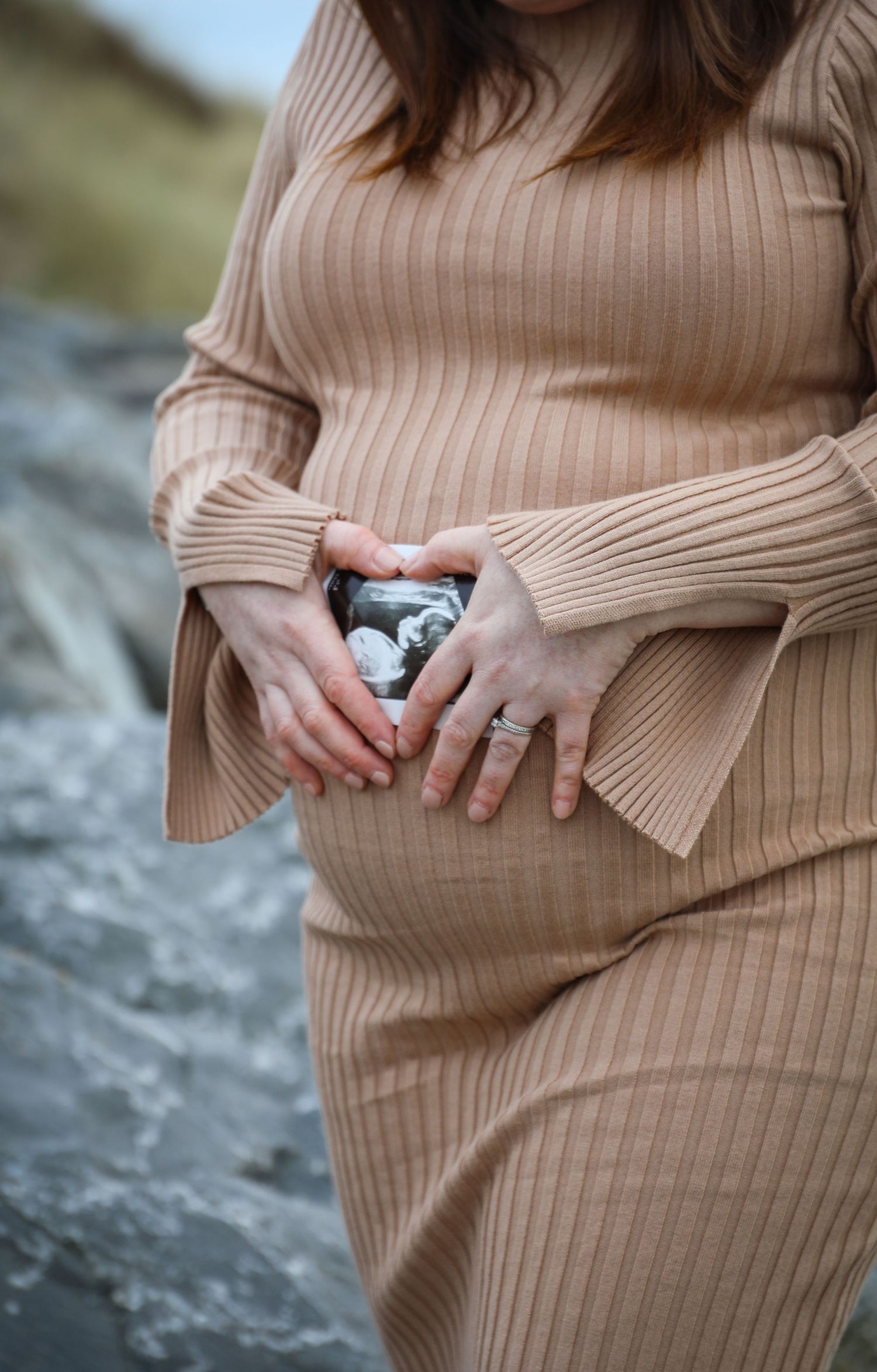 Close-up of hands holding a sonogram photo while wearing a ribbed beige sweater dress.