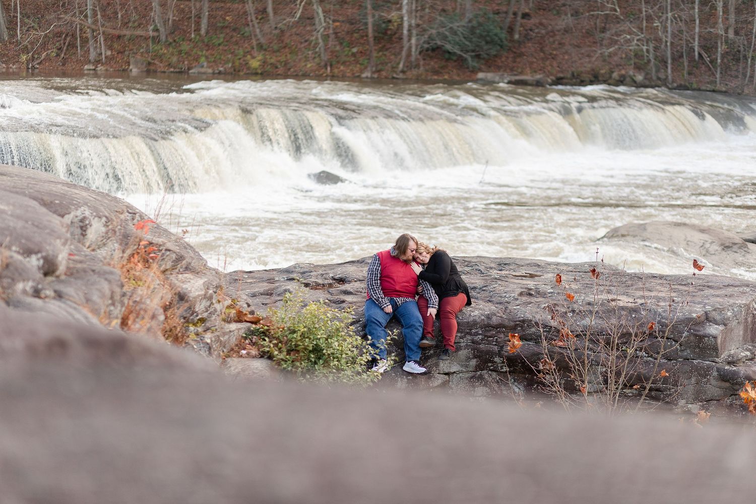Two people share a quiet moment on rocks above a scenic waterfall.
