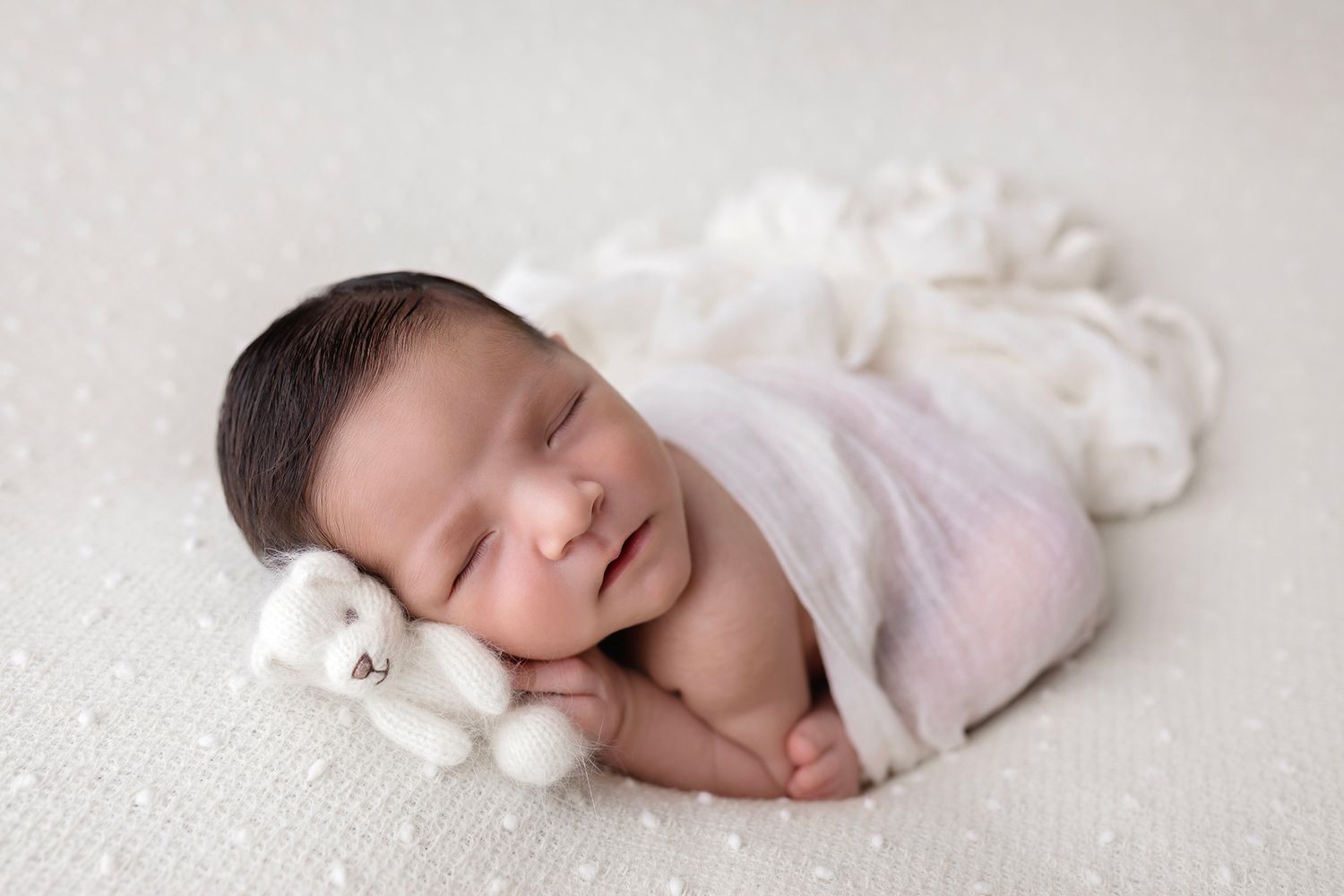 Dreaming newborn nestled with small white teddy bear on textured white surface.